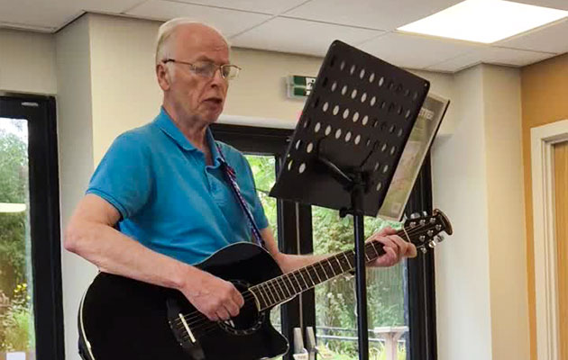 Headway Shropshire client Stephen standing indoors, playing a black acoustic guitar beside a music stand.